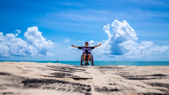 Mężczyzna na wózku inwalidzkim na plaży z radośnie uniesionymi do góry rękami 